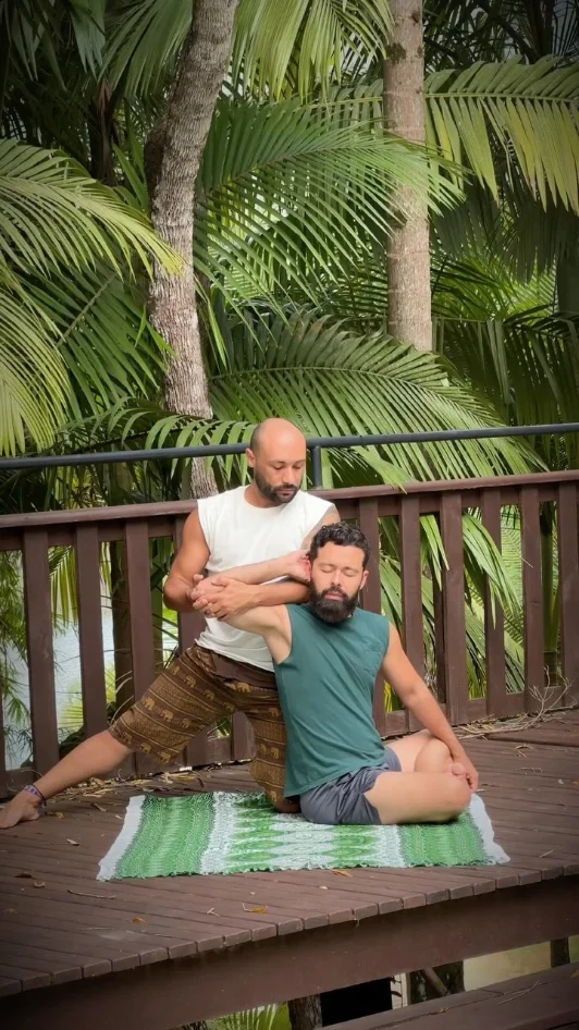 Ju aplicando um alongamento de ombros em um praticante sentado, num deck de madeira cercado de palmeiras.
